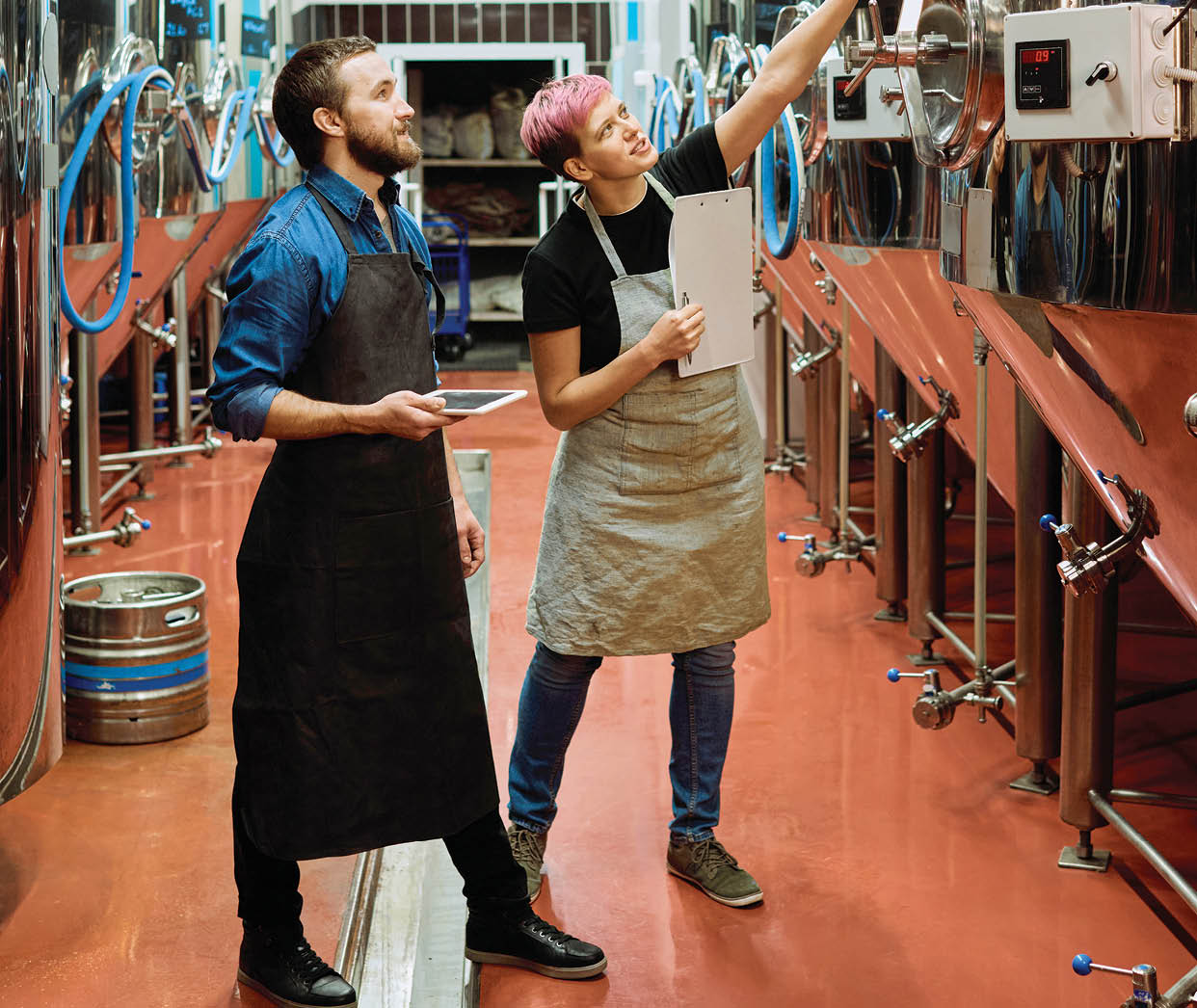 Two young workers of large beer production factory discussing new equipment while woman with clipboard pointing at huge steel tanks