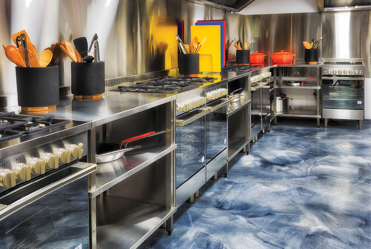 Modern brand new fully equipped kitchen with a line of ovens, cup boards, tables and utensils along the brushed aluminium walls ready to cook.